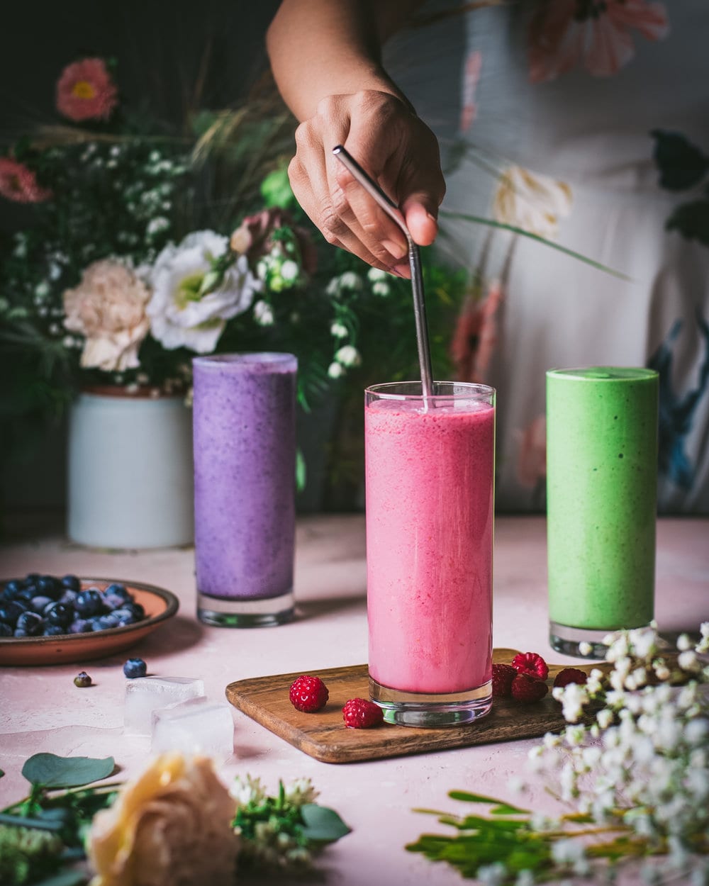 Woman stirring raspberry tahini smoothie next to flowers and two other smoothies on a table.