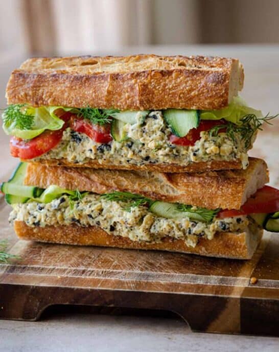 two baguette sandwiches stuffed with vegan tuna salad, cucumber, lettuce and tomato on wooden board.