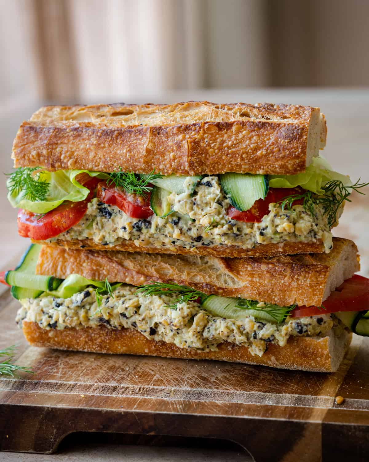 two baguette sandwiches stuffed with vegan tuna salad, cucumber, lettuce and tomato on wooden board.