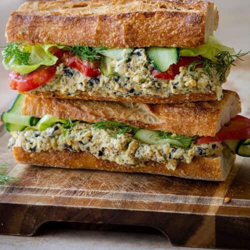 two baguette sandwiches stuffed with vegan tuna salad, cucumber, lettuce and tomato on wooden board.