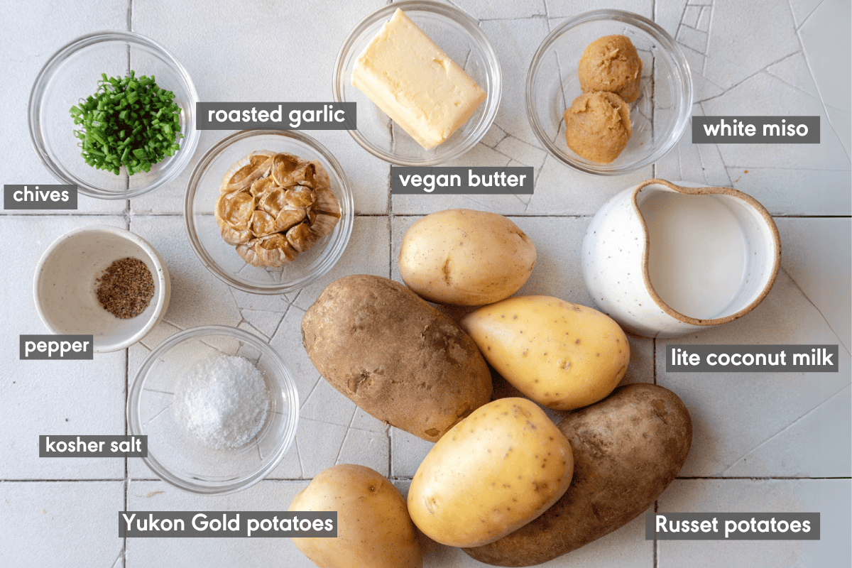 Ingredients for vegan mashed potatoes laid out on a white tiled surface with ingredients labeled. 