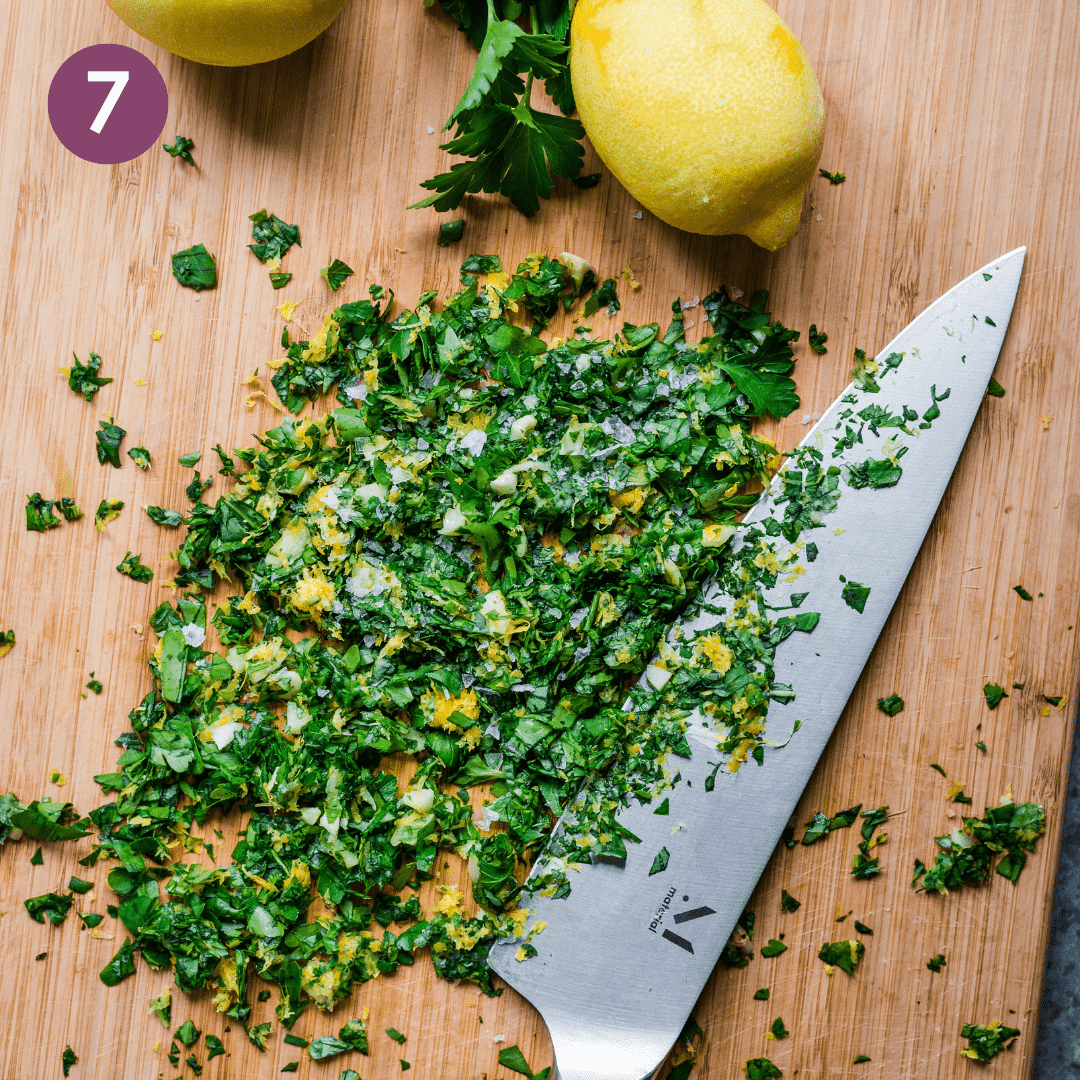 Italian gremolata chopped up with lemon zest on a wooden cutting board and zested whole lemons.