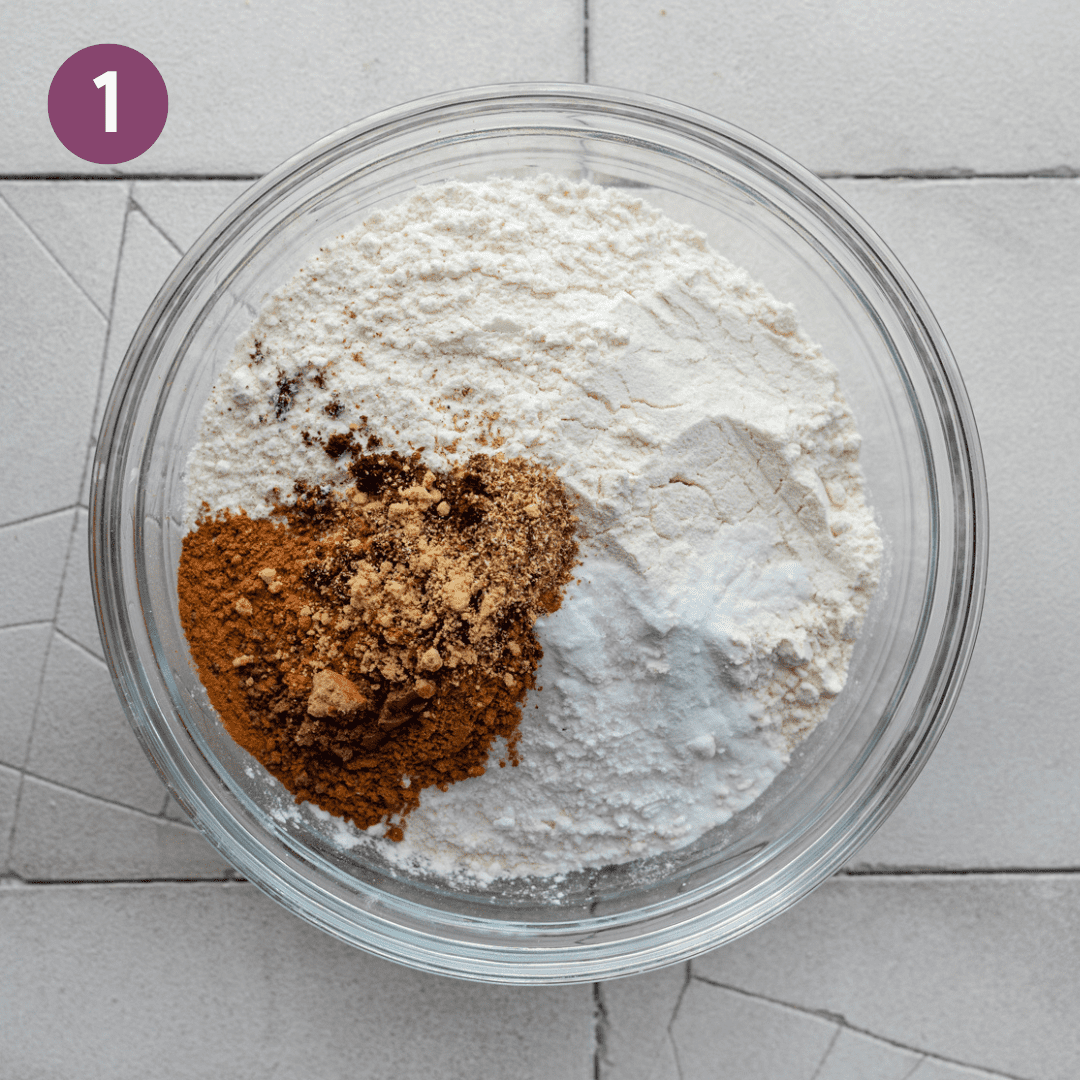 flour, baking powder, salt, and pumkin spices in a large glass bowl on white tiled surface.