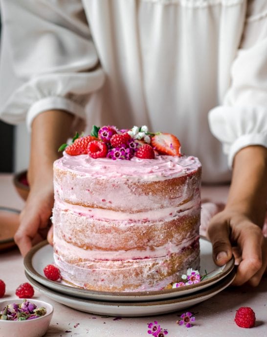 woman wearing white dressing putting down a layered pink cake topped with raspberries.