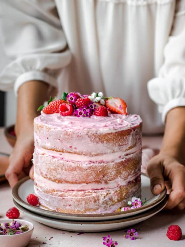 woman wearing white dressing putting down a layered pink cake topped with raspberries.