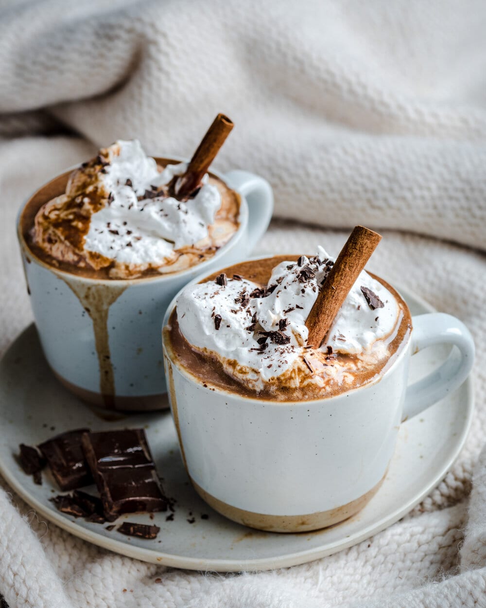 Side view of of hot chocolate in two white mugs on a white plate with chocolate squares on it.