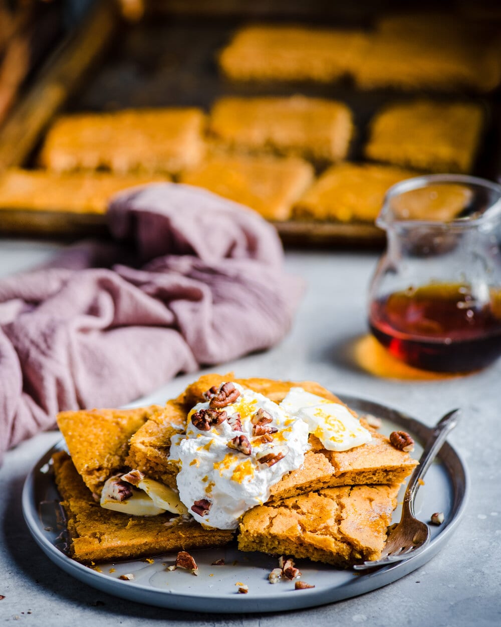 Stack of sheet pancakes on a plate next to a jar of maple syrup on a table.