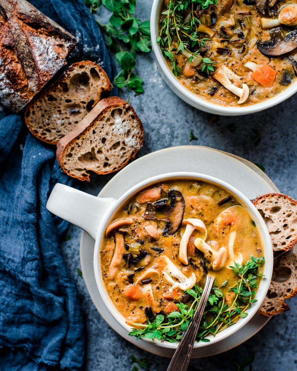 Two bowls of mushroom soup, slices of bread and a linen napkin on a blue-grey table.