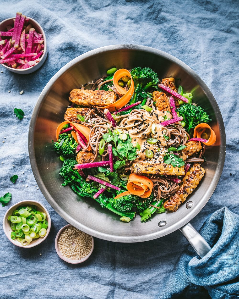 Soba noodle salad in a large frying pan on a blue tablecloth next to small bowls of ingredients.