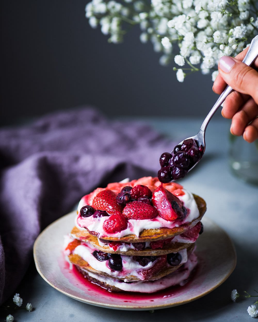Hand spooning blueberries onto stack of pancakes with yogurt and berries on a grey plate.
