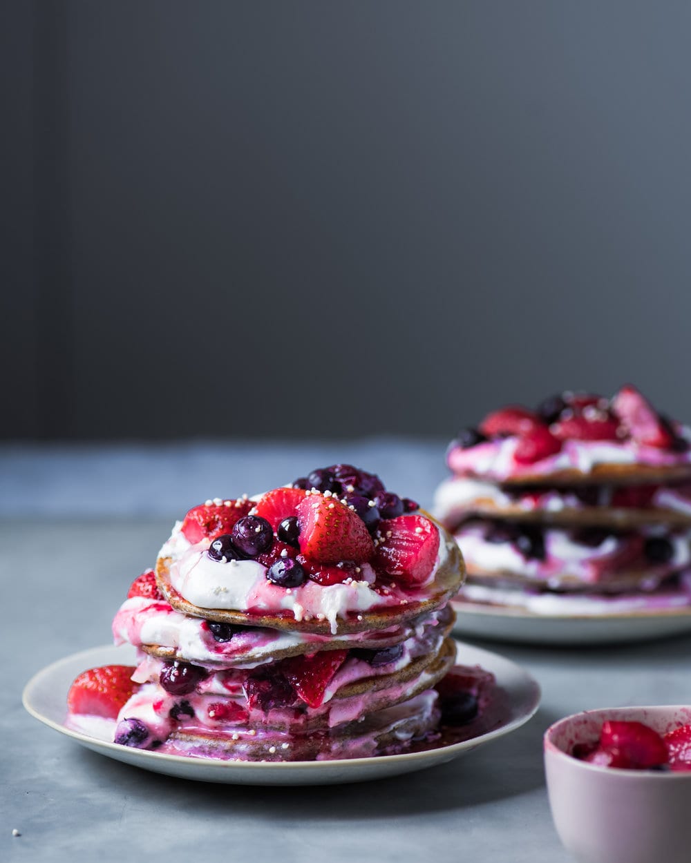 Two grey plates stacked high with pancakes layered with vegan yogurt and berries.