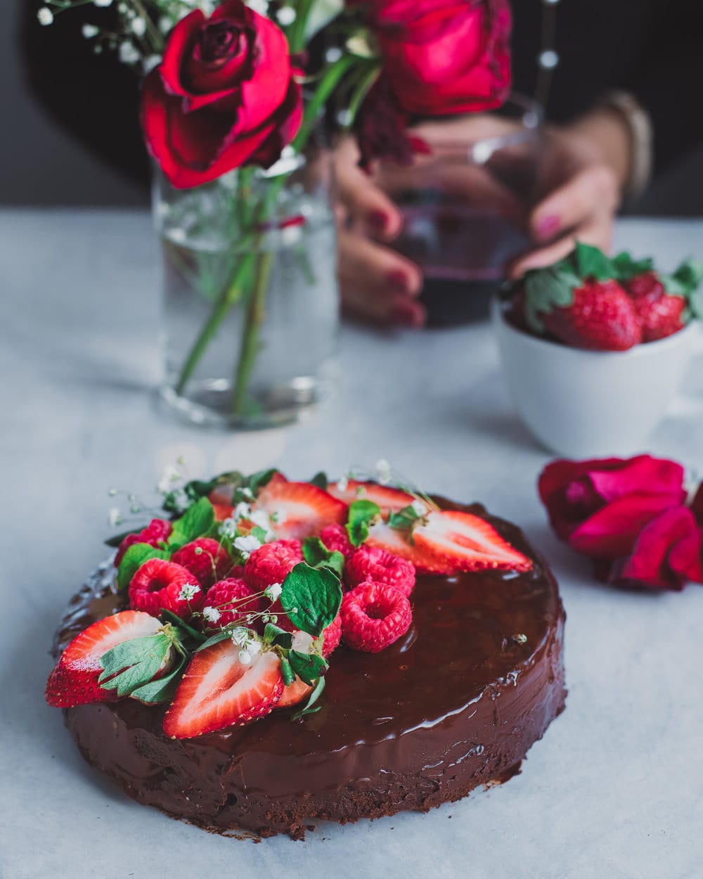 Chocolate cake with berries on white table next to roses and bowl of berries.