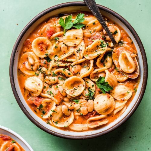 italian pasta and white bean stew in a ceramic bowl on a green surface.