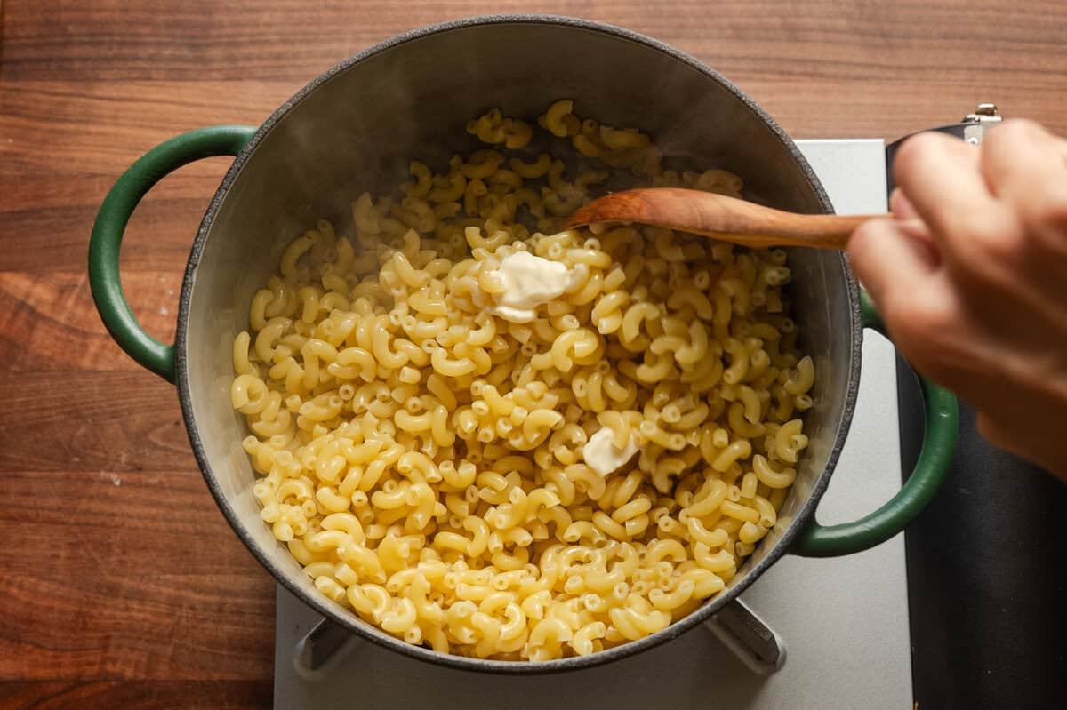 wooden spoon stirring butter into cooked macaroni in a green saucepan.