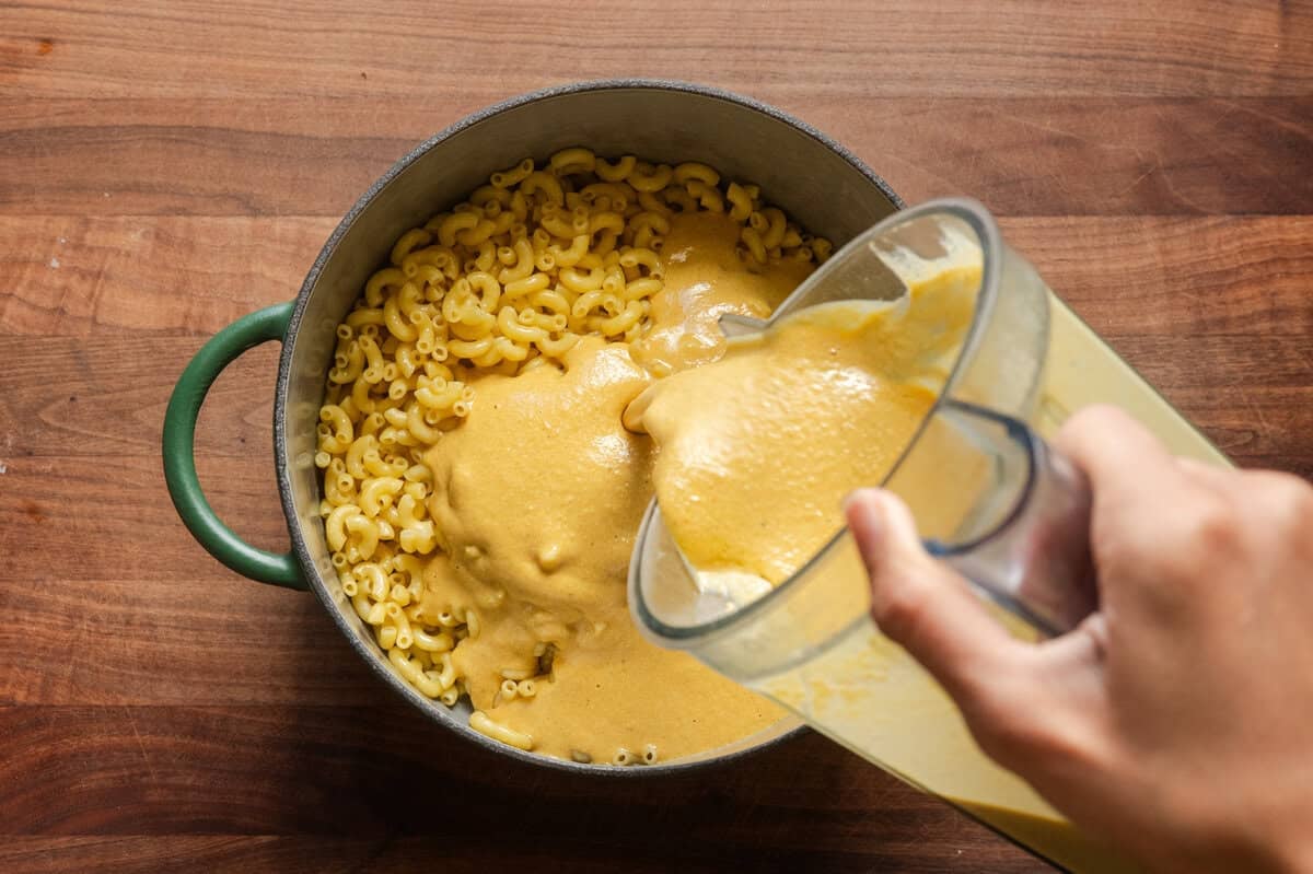 pouring vegan cheese sauce from a blender into cooked macaroni in a green saucepan.