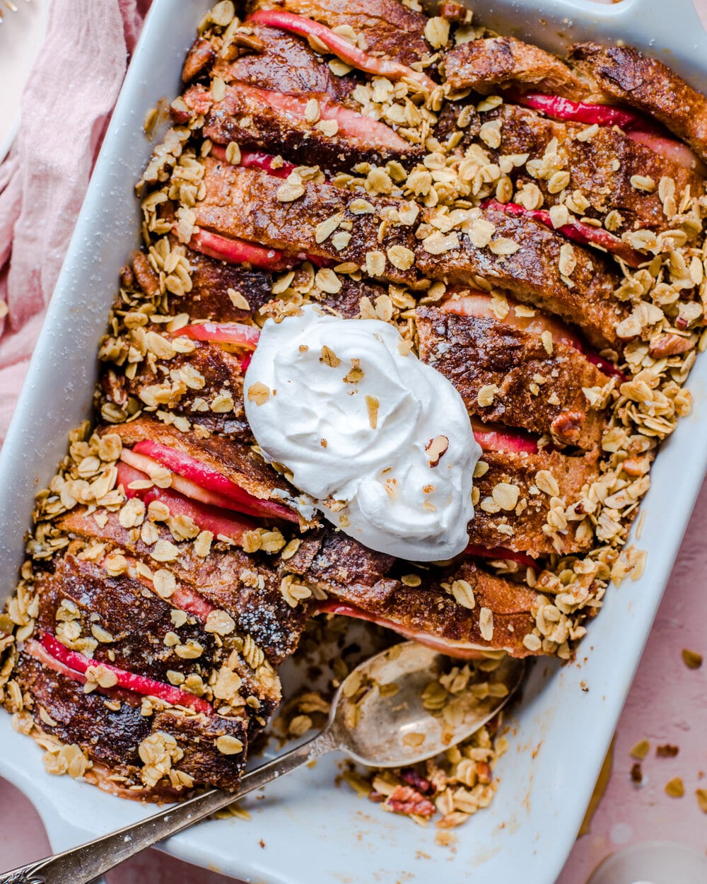 French Toast Casserole with whipped cream in a white casserole dish on a pink table.