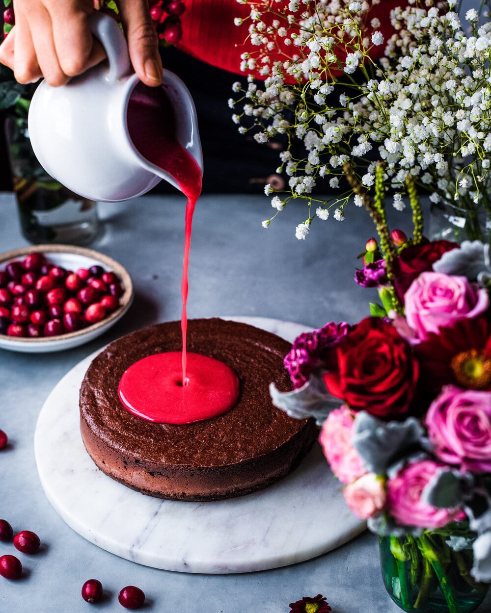 Person pouring cranberry-raspberry compote onto cheesecake on marble serving board.