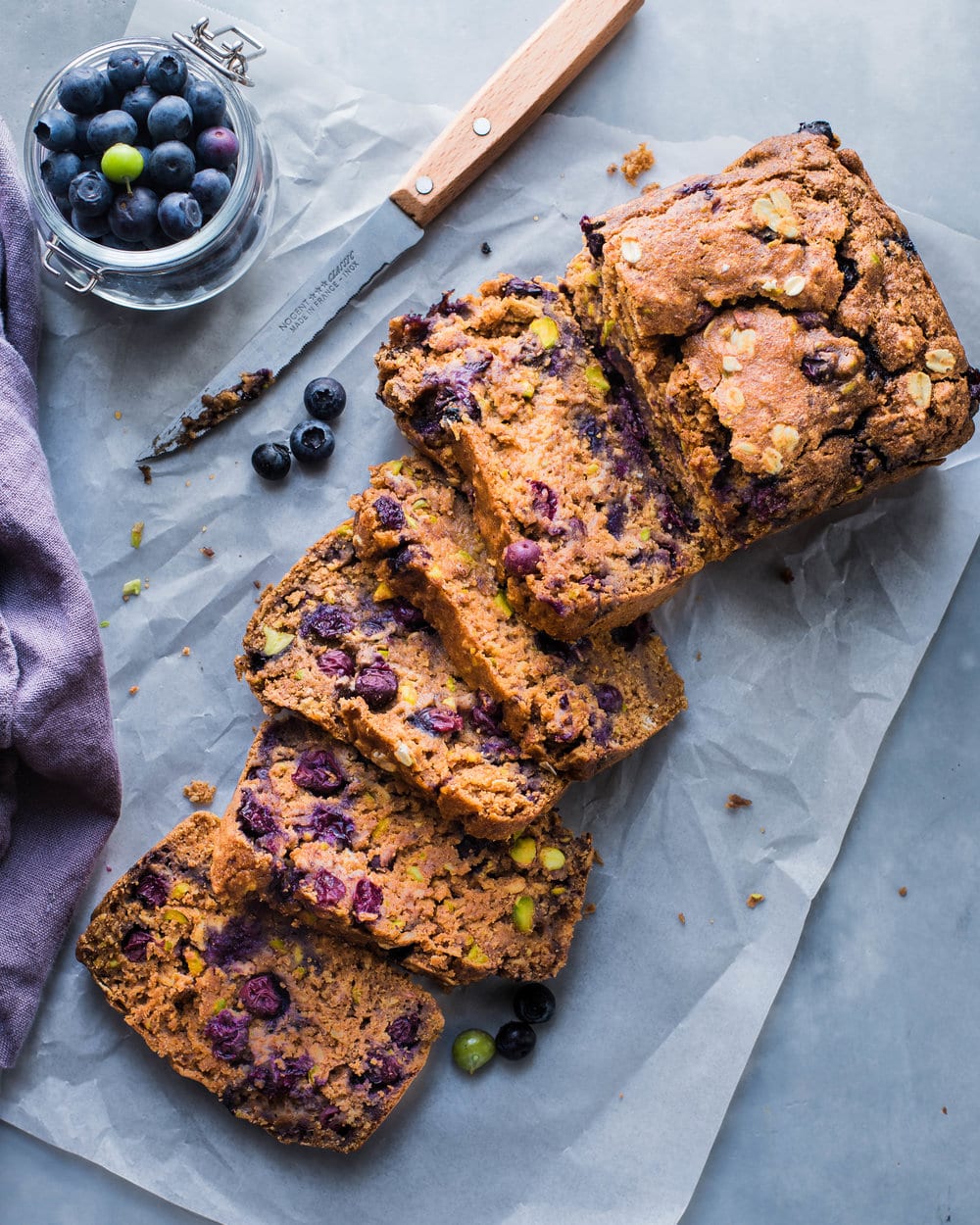 Sliced blueberry breakfast cake on parchment paper next to a knife and jar of blueberries.