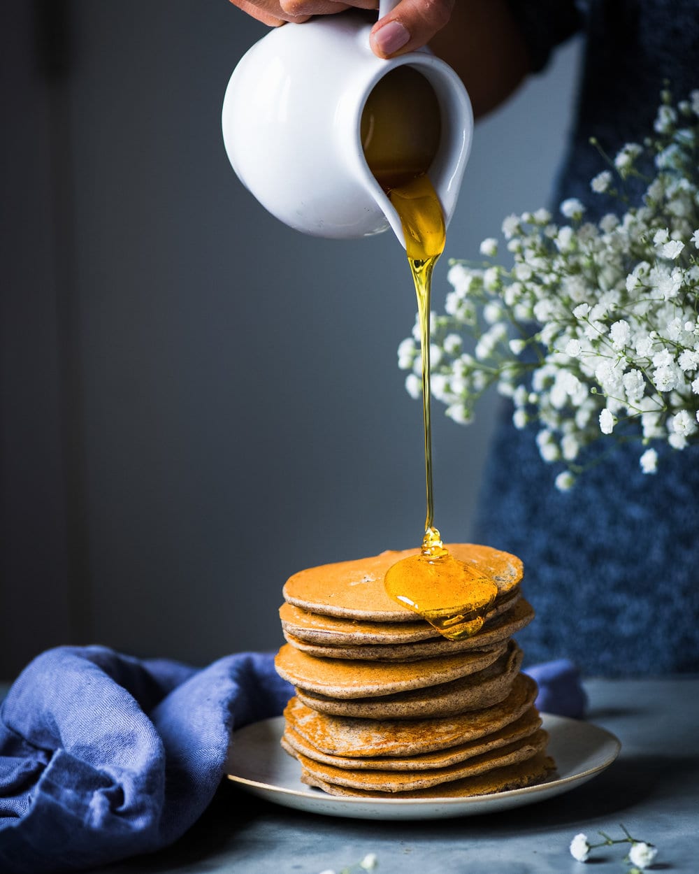 Side view of maple syrup being poured onto tall stack of pancakes on a grey plate.