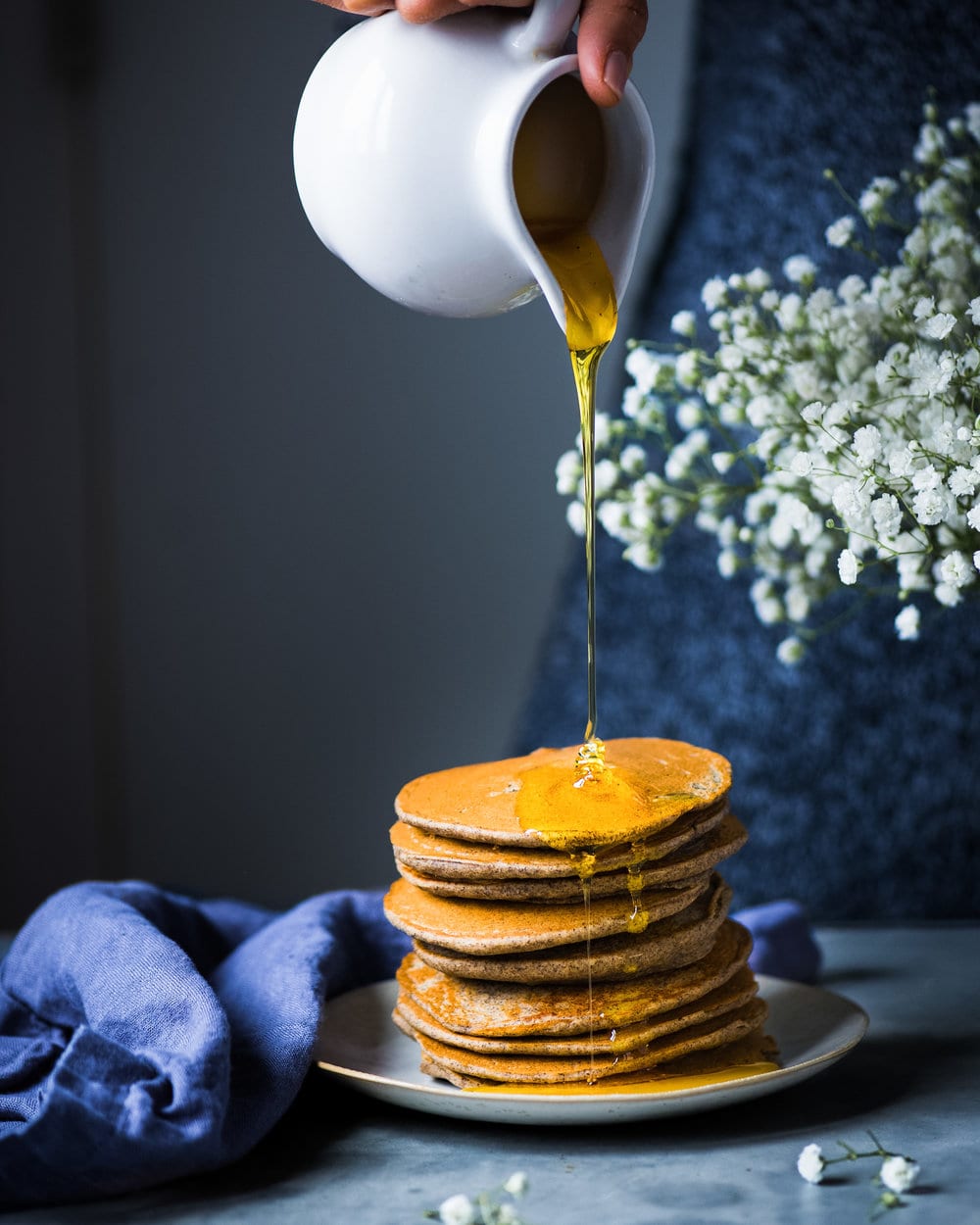 Side view of maple syrup being poured onto tall stack of pancakes on a grey plate.