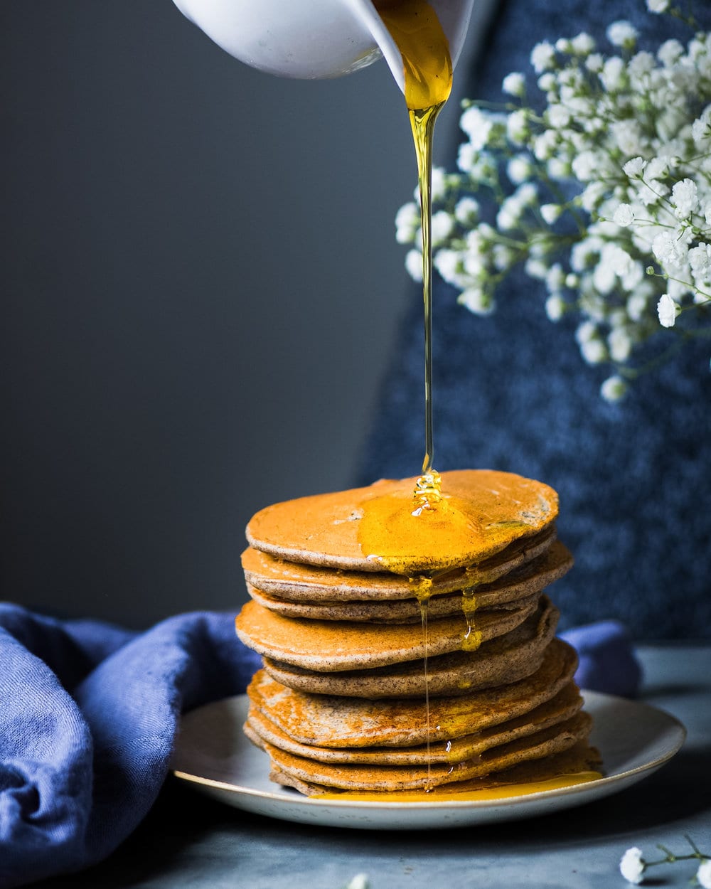 Maple syrup being poured onto tall stack of pancakes on a grey plate.