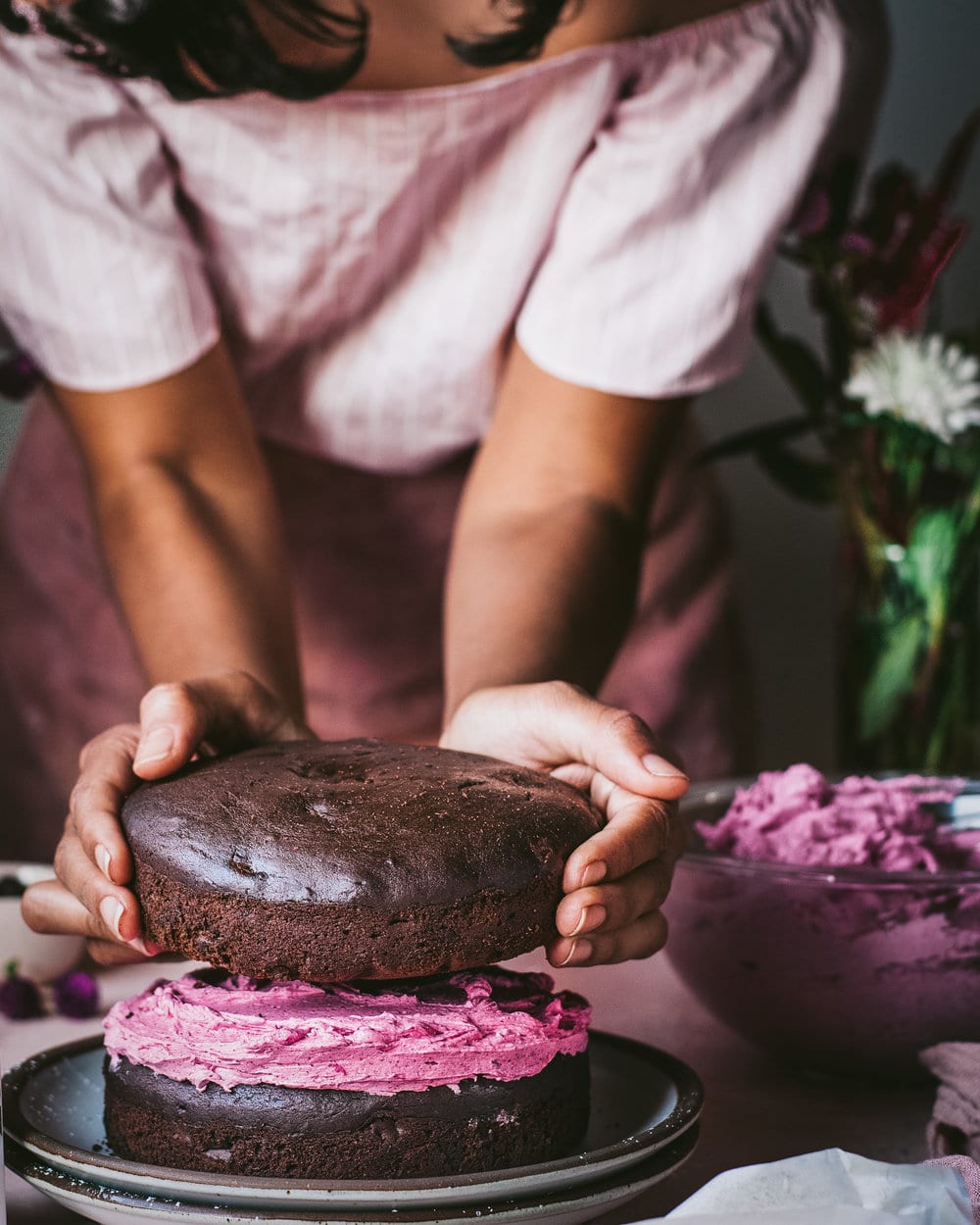 Woman placing second layer of cake onto first layer with frosting.