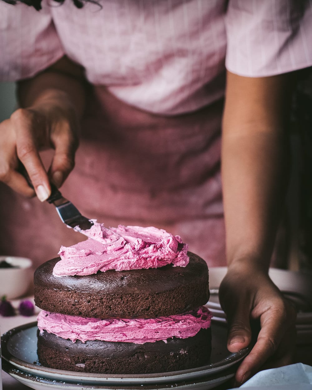 Woman smoothing frosting onto top of cake sitting on two stacked plates on a table.