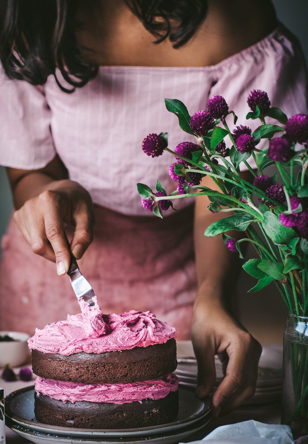 Woman smoothing hibiscus frosting onto chocolate layer cake.
