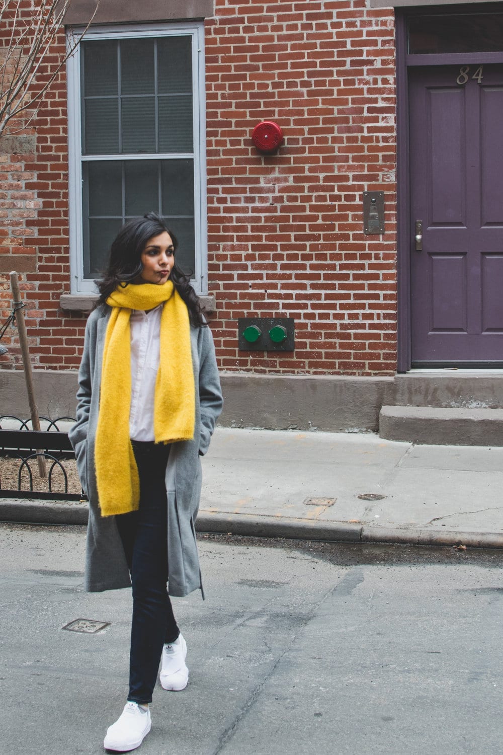 Nisha Vora wearing a bright yellow scarf and grey long sweater crossing the street in front of a brick building.