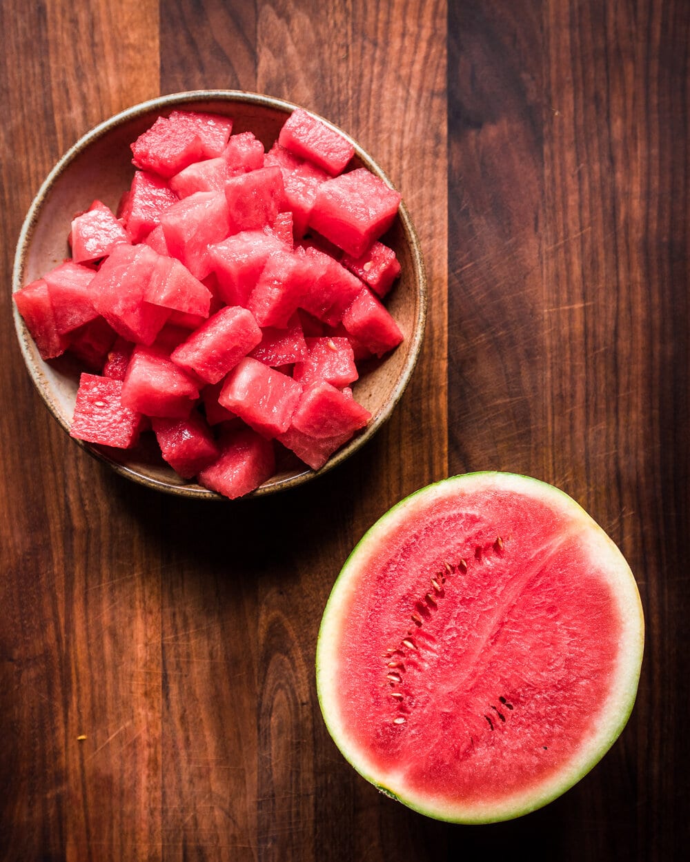 Half a watermelon and a bowl of diced watermelon on a wooden table.