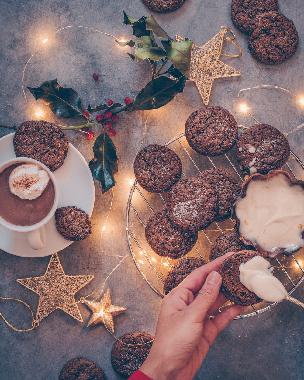 Woman frosting gingerbread cookies next to holiday decorations and lights on a table.