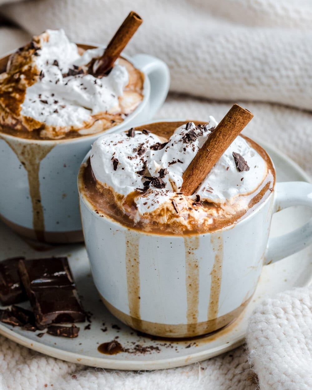 Side view of hot chocolate in two white mugs on a white plate with chocolate squares on it.