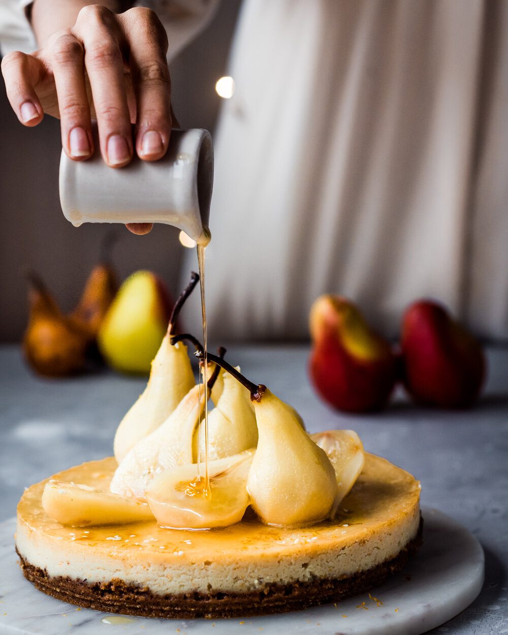 Woman pouring poaching syrup onto baked cheesecake topped with pear halves.