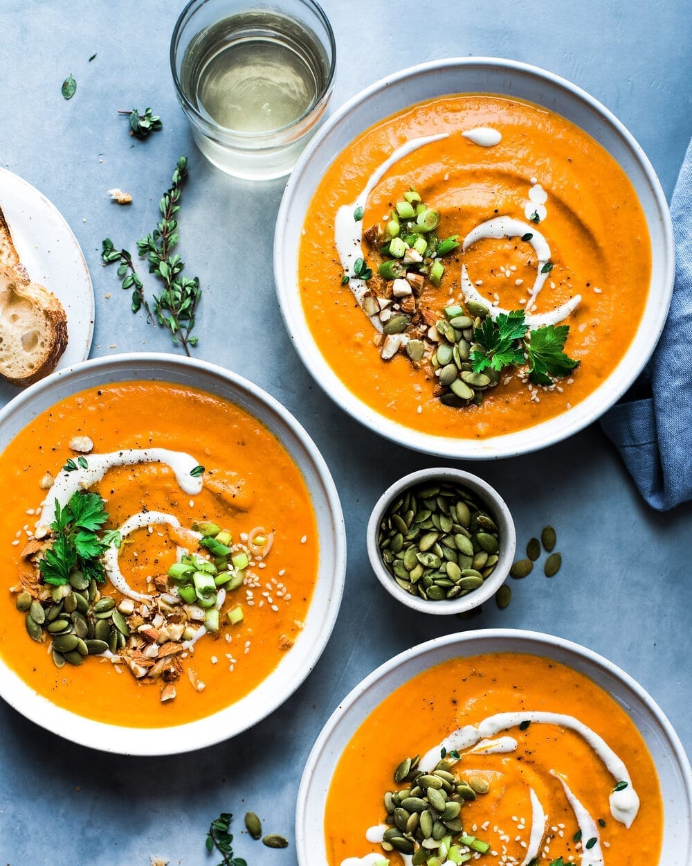 Three bowls of soup and a small bowl of pumpkin seeds on a grey table.