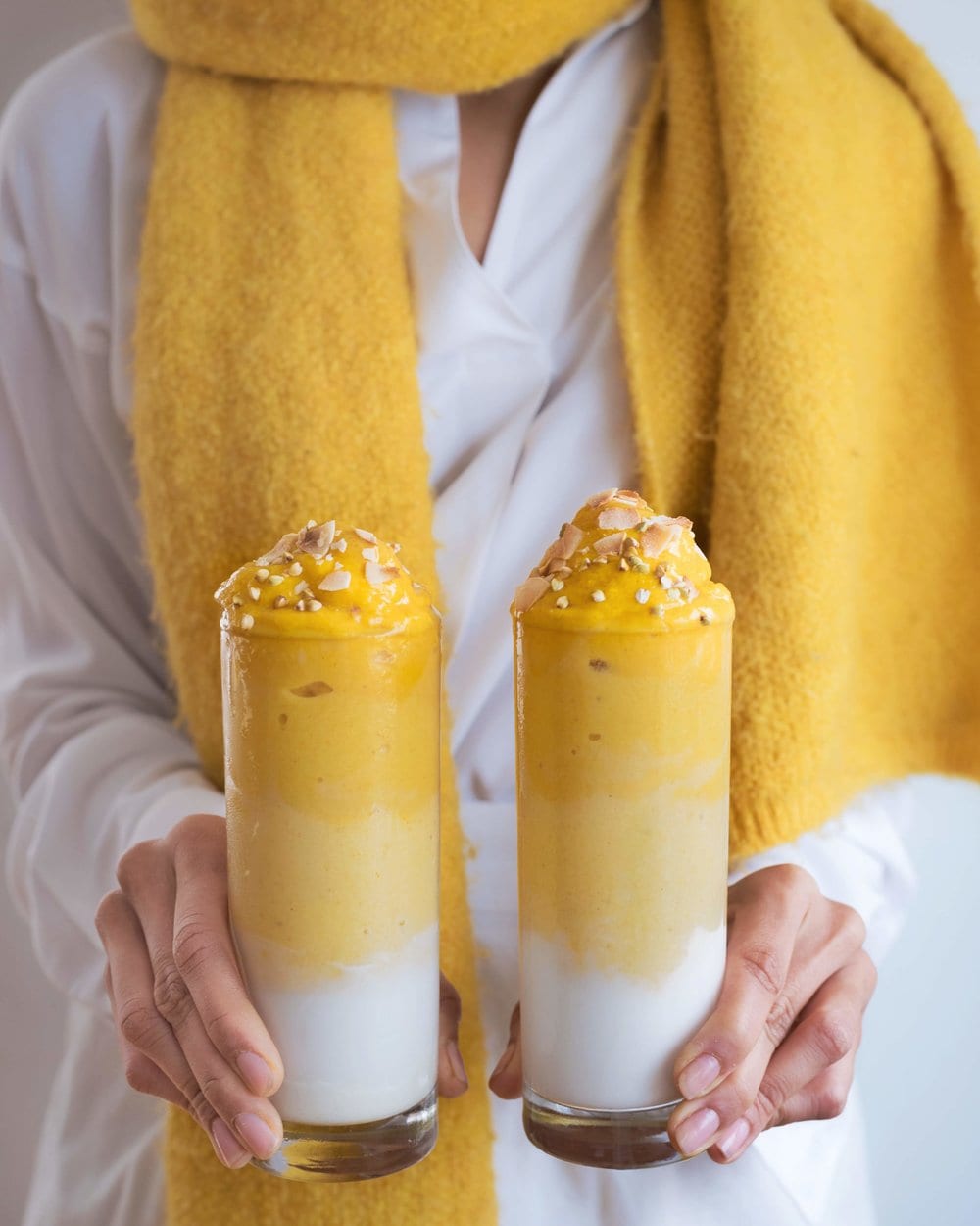 Woman wearing yellow scarf holding two white and yellow mango smoothies.