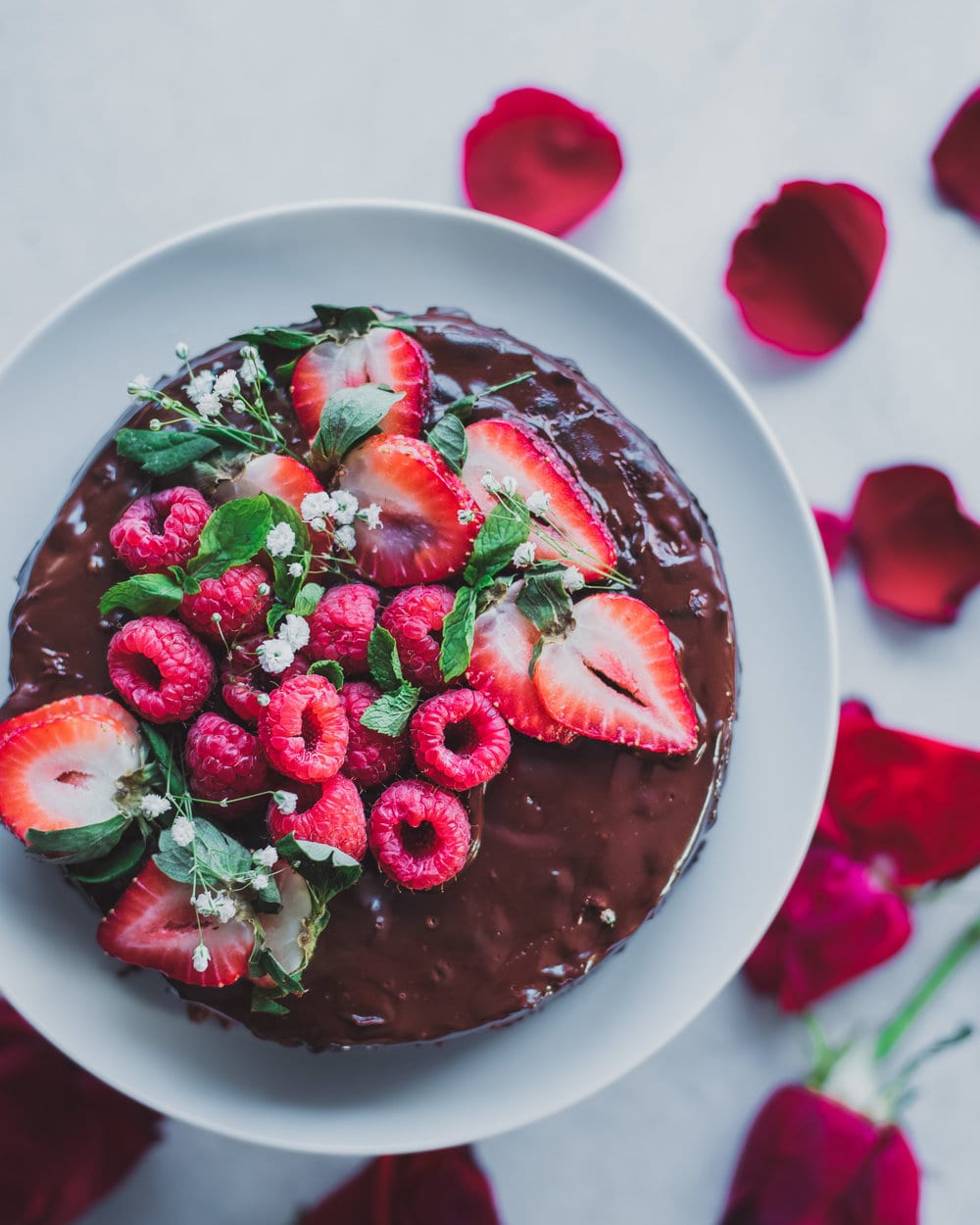 Chocolate caked topped with berries on a white plate next to rose petals.