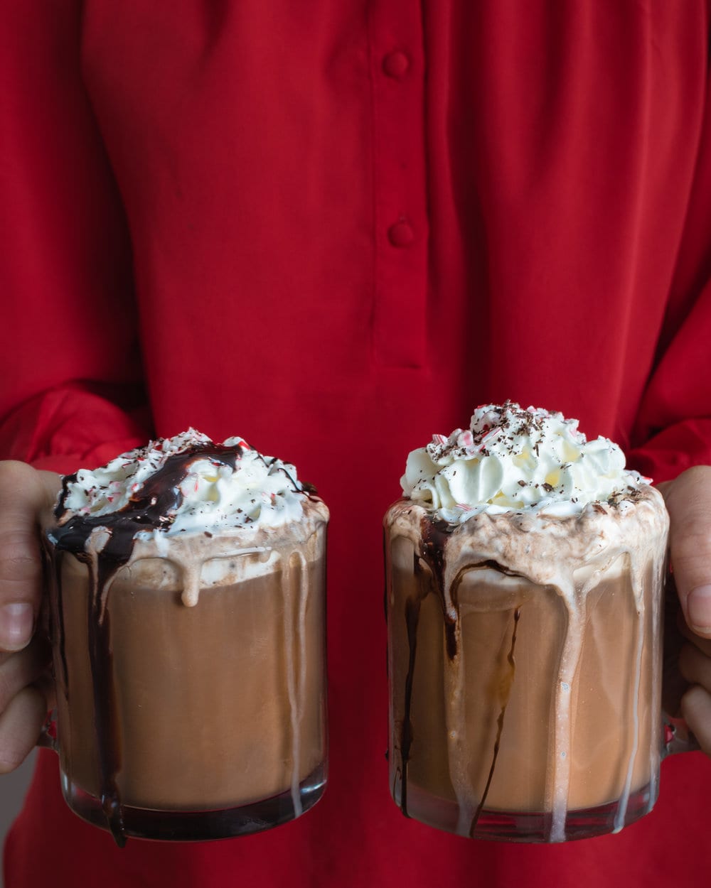 Person wearing red holding two mugs filled with peppermint mocha and whipped cream.