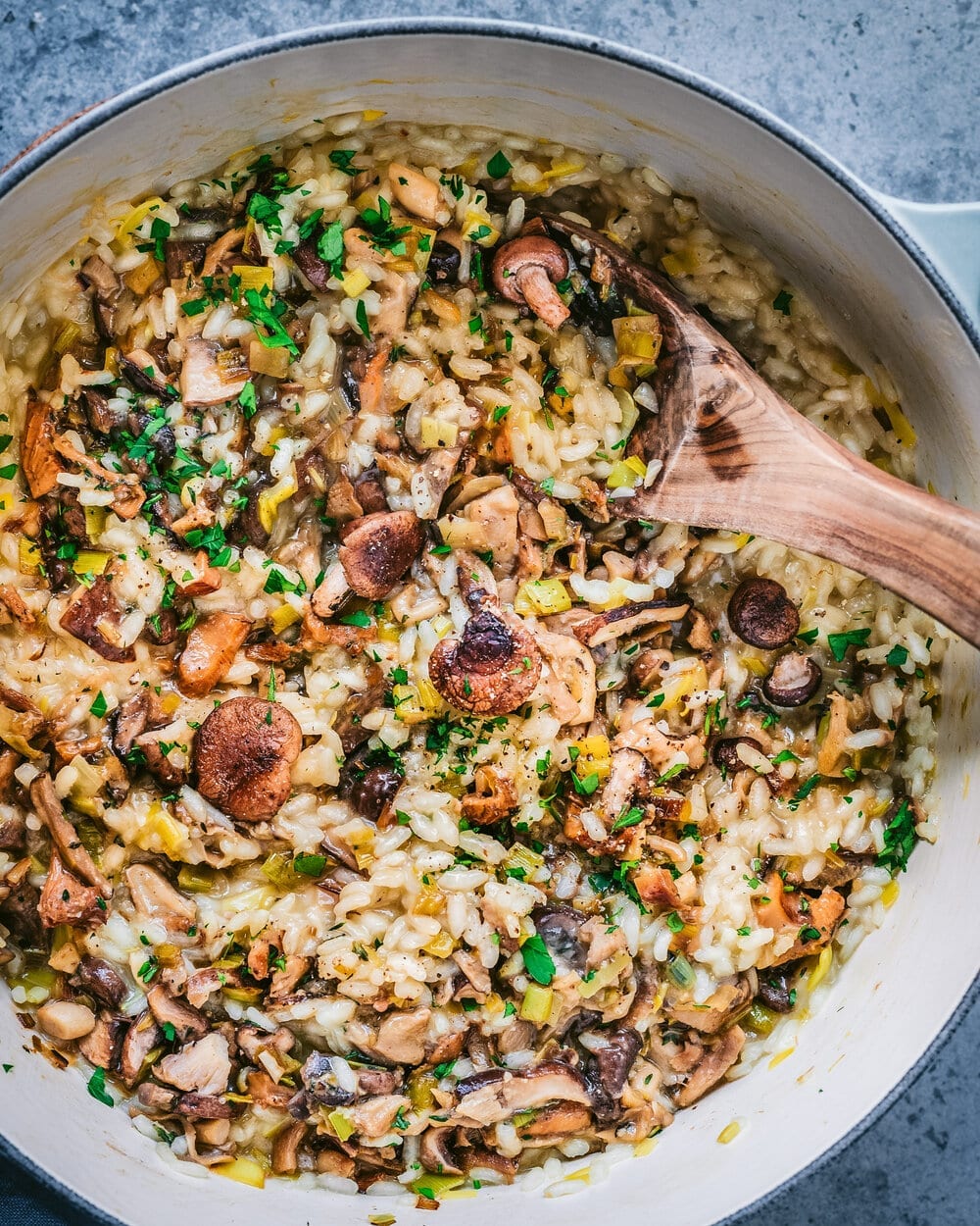 a blue dutch oven filled with vegan risotto with mushrooms with a wooden spoon dug into the risotto. 