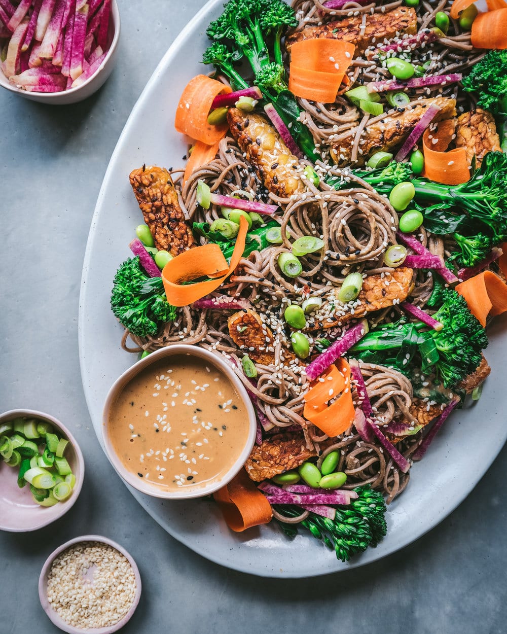 Soba noodle salad on a large white platter with a bowl of sauce on a grey table.