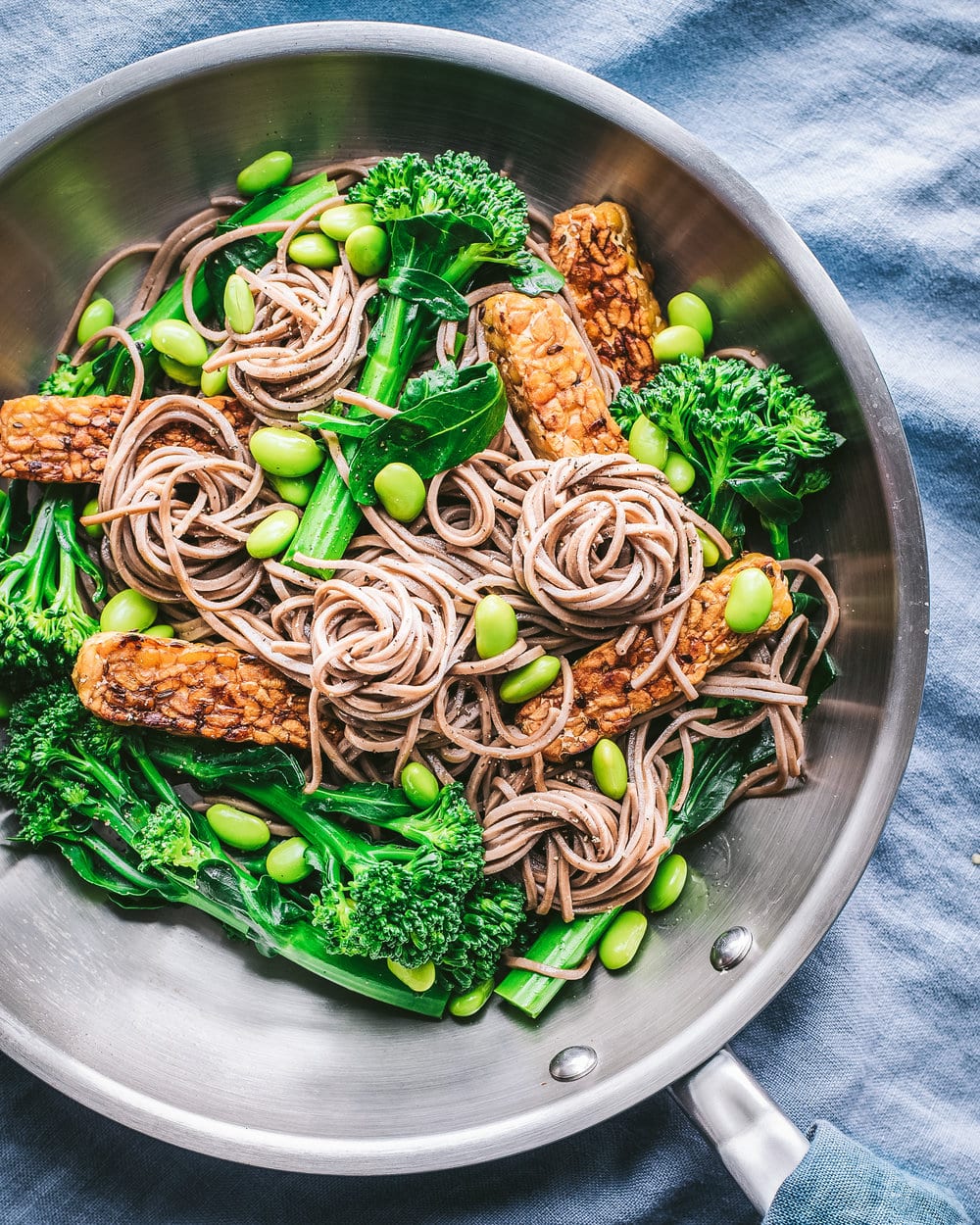 Tempeh, edamame, noodles and broccolini in a large frying pan on a blue table cloth.