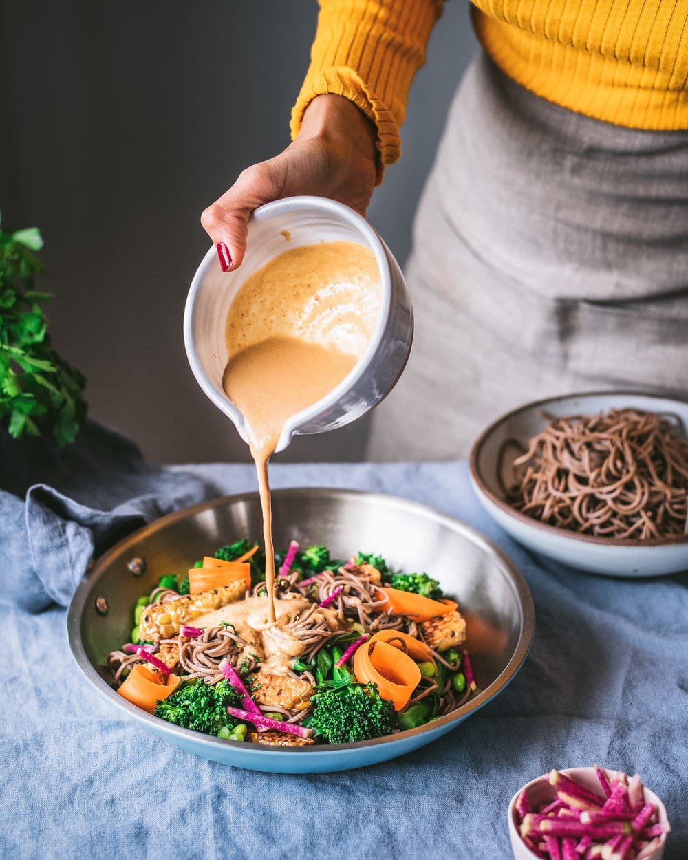 Woman pouring sesame-cashew sauce onto soba noodle salad on a table.