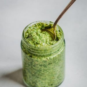 cilantro pesto in a glass jar with a spoon on white backdrop.