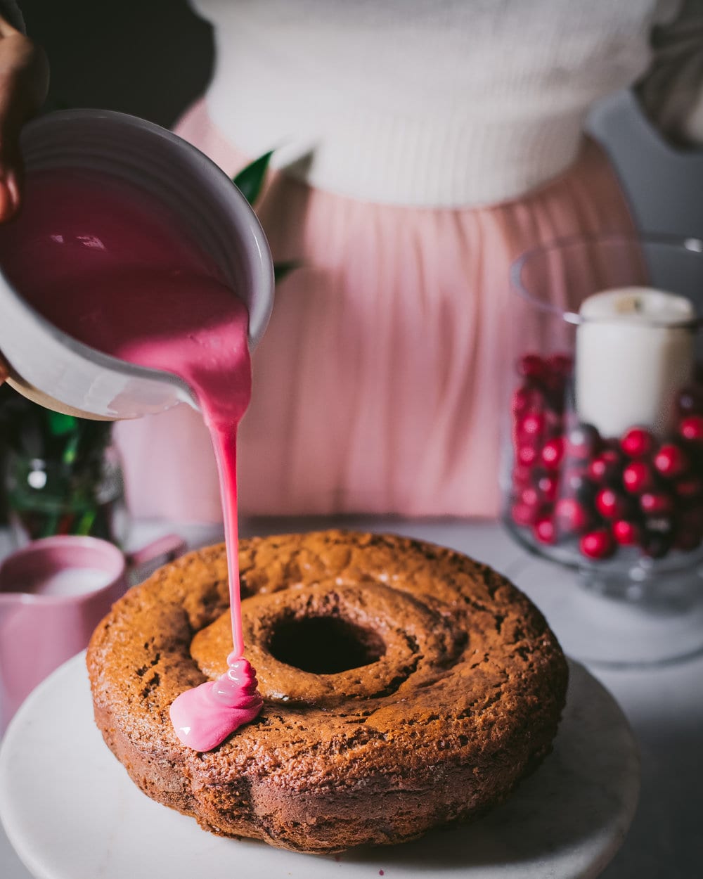 Woman pouring pomegranate glaze from a small bowl onto bundt cake on a cake stand.
