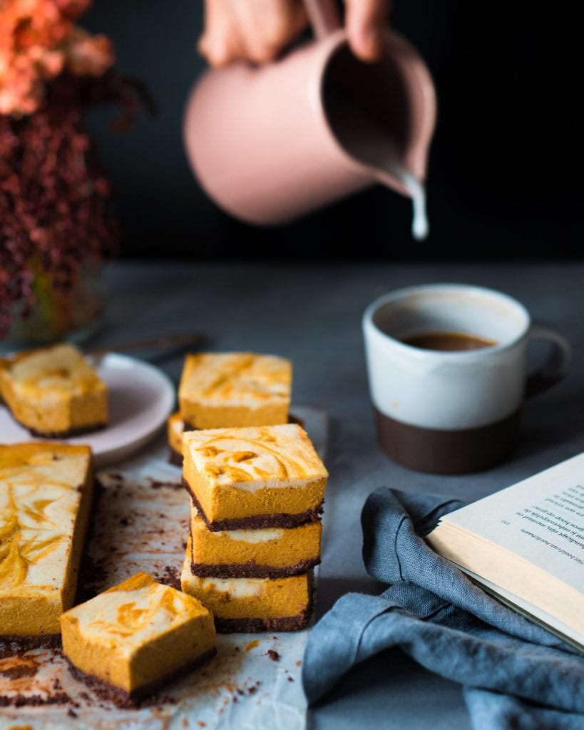 Woman pouring vegan milk into a cup of coffee next to cheesecake bars on a table.