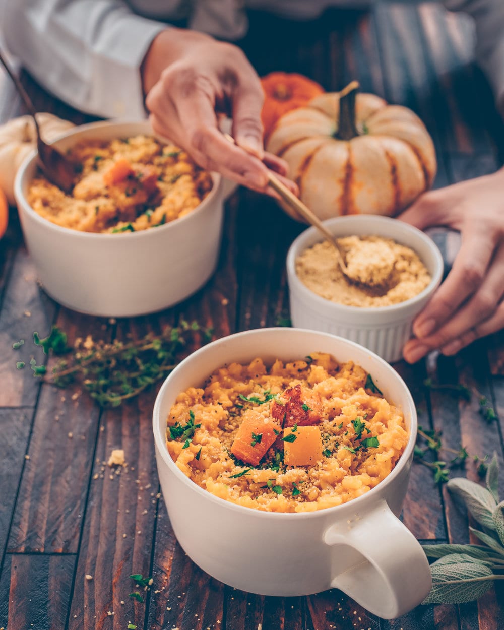 Woman spooning vegan parmesan onto two mugs of pumpkin risotto on a wood table.