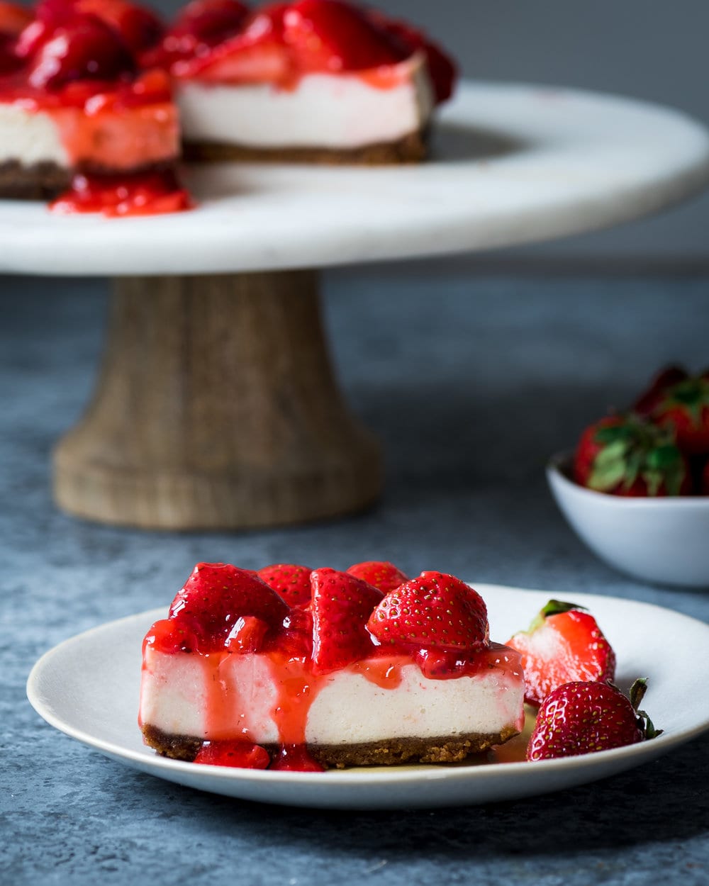 Slice of cheesecake with strawberries on a plate in front of rest of cheesecake on a stand.