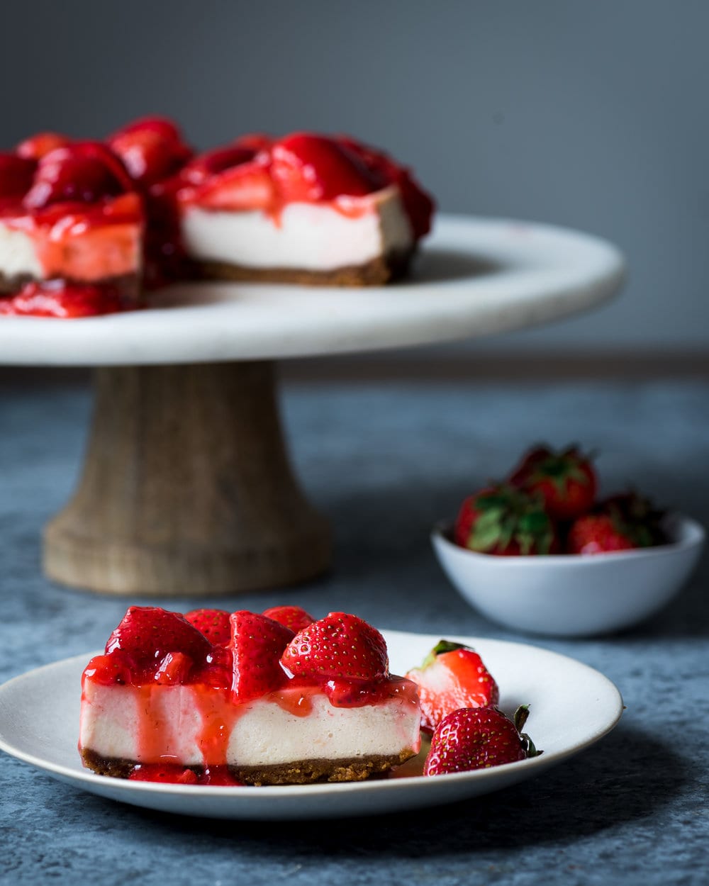 Slice of cheesecake with strawberries on a plate in front of rest of cheesecake on a stand.