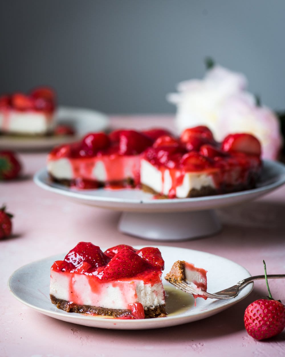 Vegan cheesecake slice covered in strawberry sauce on pink table.
