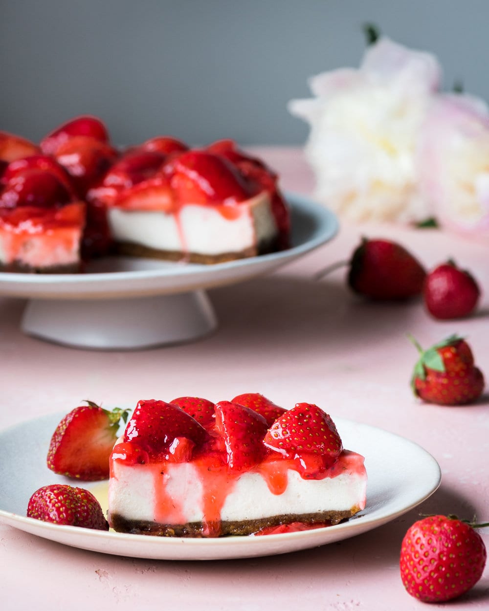 Slice of cheesecake with strawberries on a plate in front of rest of cheesecake on a stand.