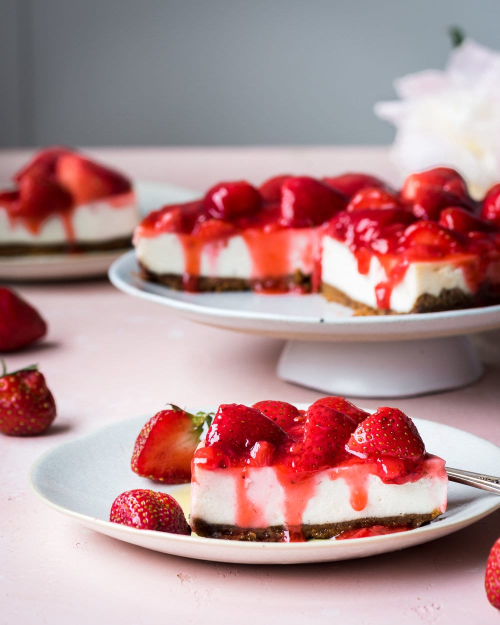 Slice of cheesecake with strawberries on a plate in front of rest of cheesecake on a stand.